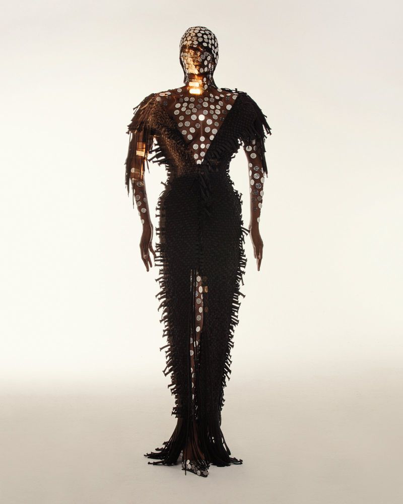 A person is wearing a black floor-length gown embellished with silver circular patterns, complete with a matching head covering against a white backdrop.