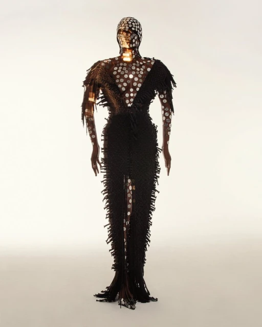 A person is wearing a black floor-length gown embellished with silver circular patterns, complete with a matching head covering against a white backdrop.