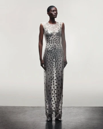 A slender Black woman poses in a sleeveless, floor-length, semi-sheer dress with circular embellishments against a plain background.