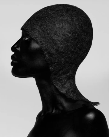 Monochromatic portrait of a Black woman in a textured black head covering against a light gray backdrop in a photo studio setting.