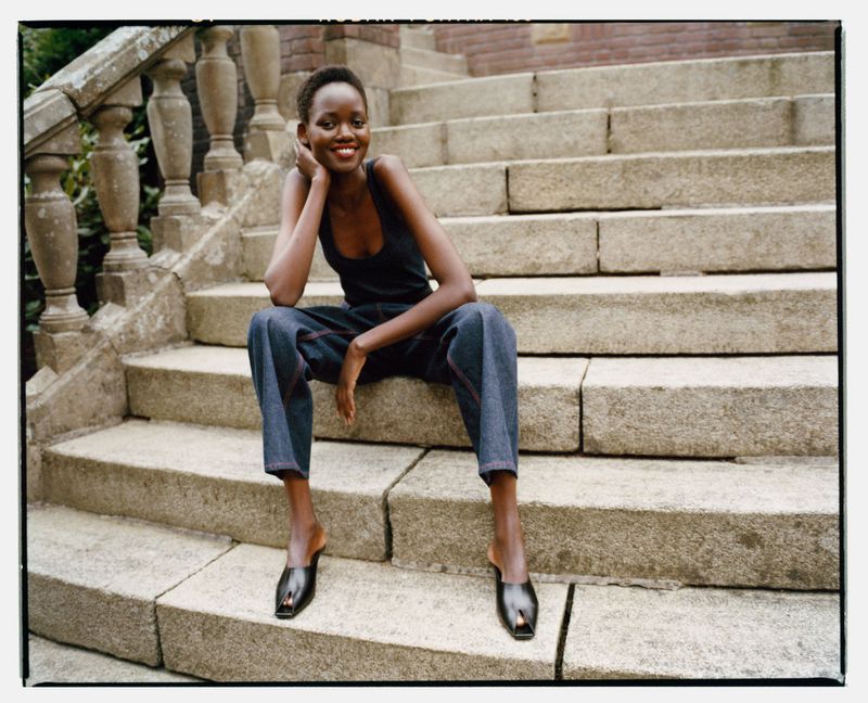 A woman sits on outdoor stone steps wearing a dark tank top, wide denim pants with red stitching, and black heeled mules.