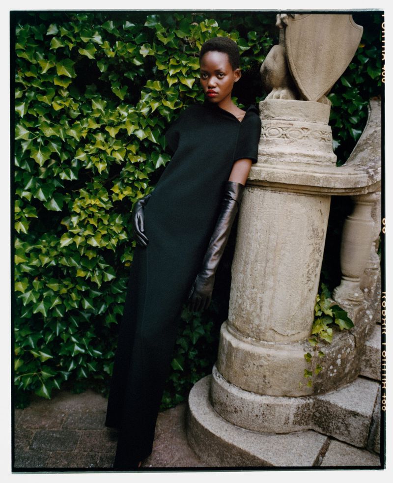 An elegant Black woman in a black dress and long gloves poses next to a stone balustrade, with ivy in the background.