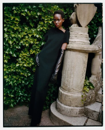 An elegant Black woman in a black dress and long gloves poses next to a stone balustrade, with ivy in the background.