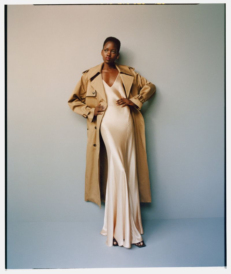 A fashionable Black woman poses in a beige trench coat over a champagne-colored silk dress against a pale blue background in a studio shot.