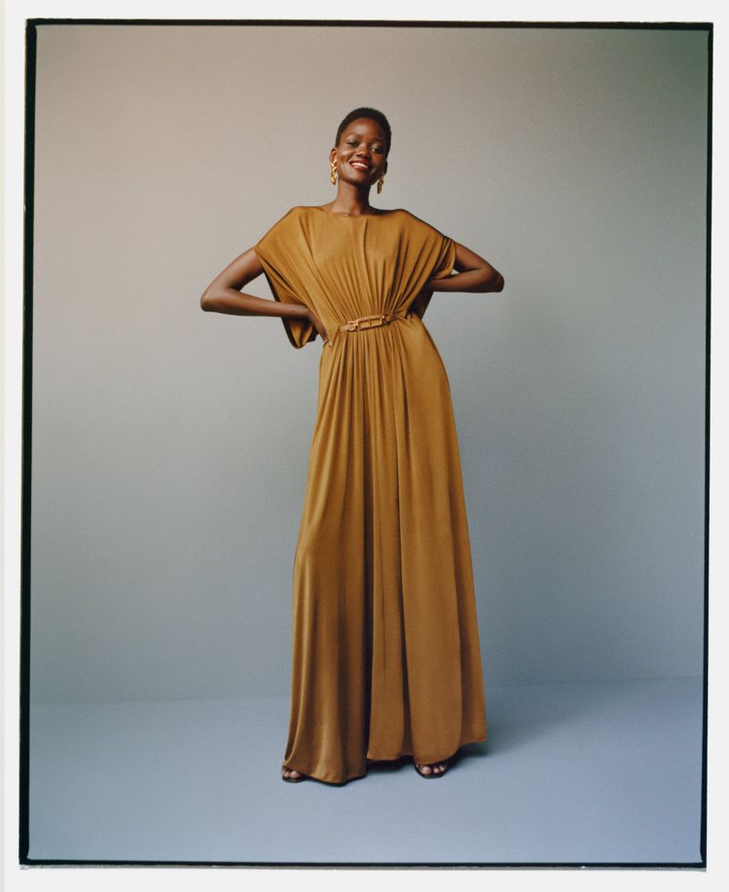 Elegant African woman in golden brown maxi dress with decorative belt posing in photo studio with neutral background. Fashion editorial.