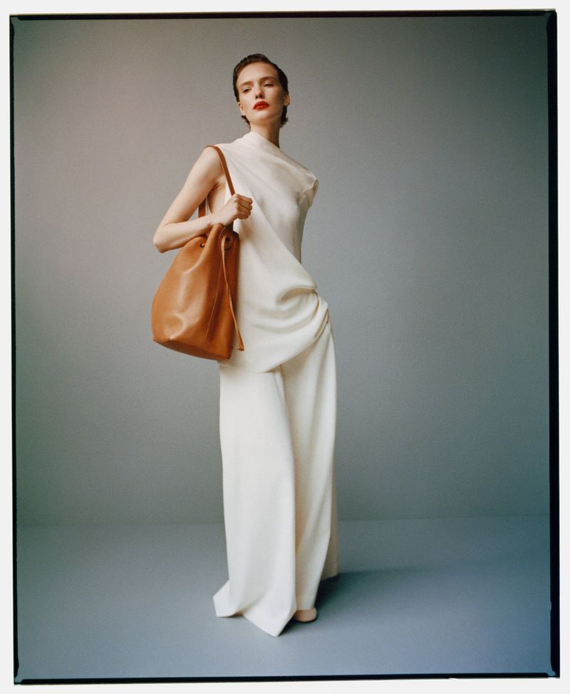 Elegant woman in a white dress and brown shoulder bag posing in a studio setting, minimalist fashion photography, soft lighting, refined style.