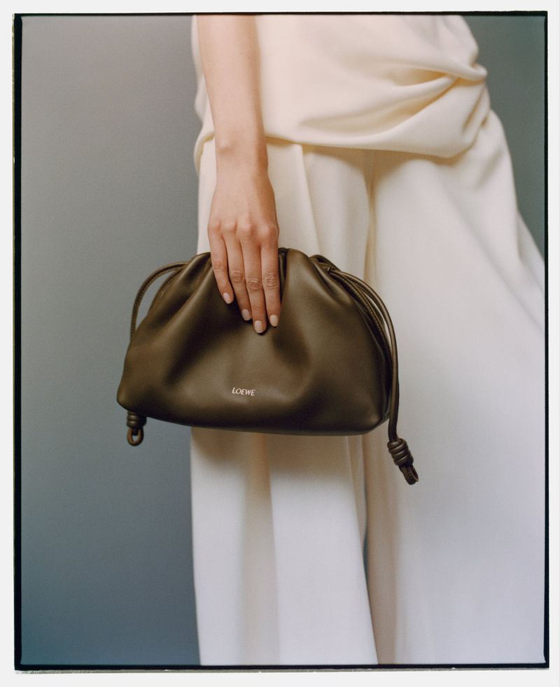 Olive-green leather pouch bag held by a fair-skinned person wearing white wide-legged pants and top against a light gray background.