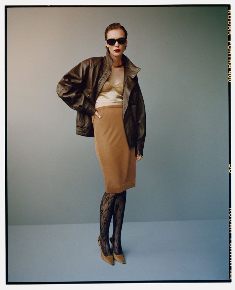 Fashionable woman in sunglasses, brown leather jacket, tan skirt, patterned stockings and heels poses in a photo studio setting.