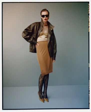 Fashionable woman in sunglasses, brown leather jacket, tan skirt, patterned stockings and heels poses in a photo studio setting.