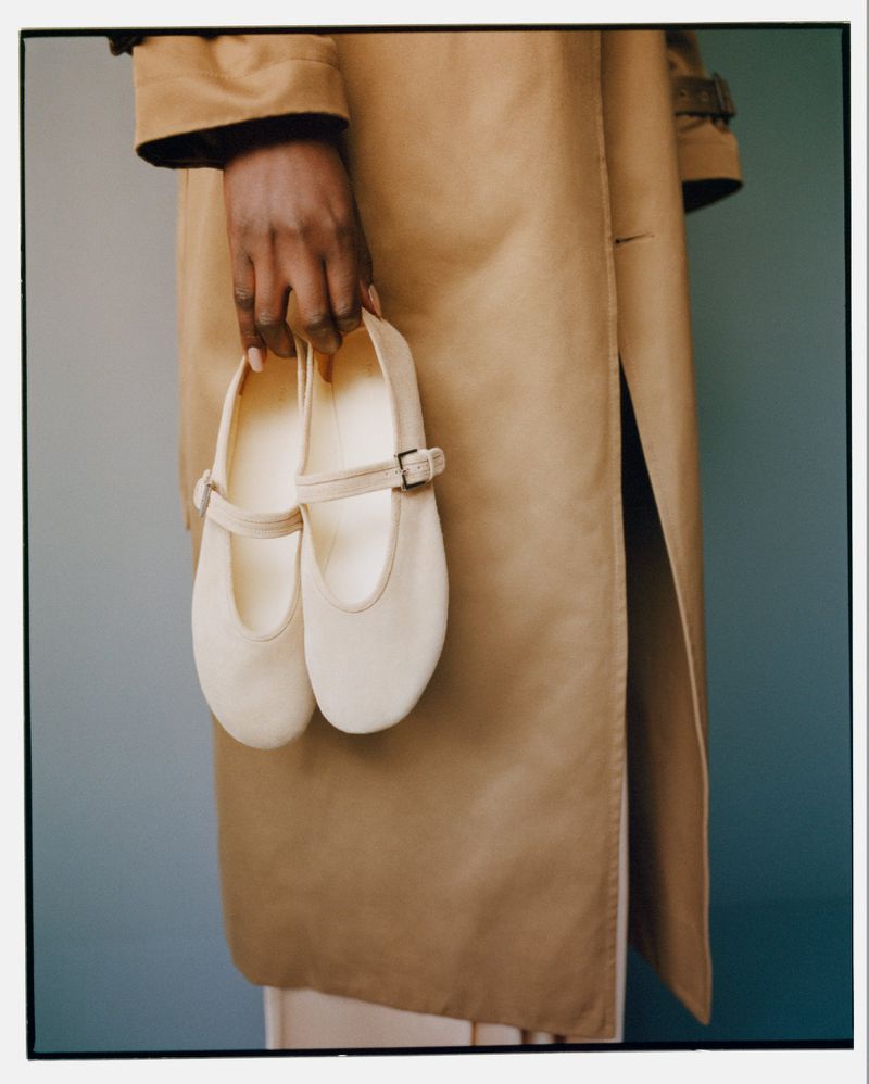 Woman holds a pair of cream-colored Mary Jane shoes in her hand, wearing a tan trench coat against a soft blue background.