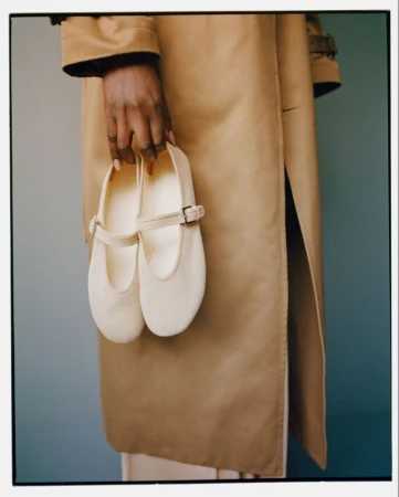 Woman holds a pair of cream-colored Mary Jane shoes in her hand, wearing a tan trench coat against a soft blue background.