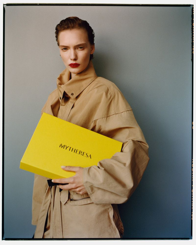 A fashionable woman in a beige jacket holds a yellow box with red lipstick against a grey backdrop in an eye-level, studio shot.