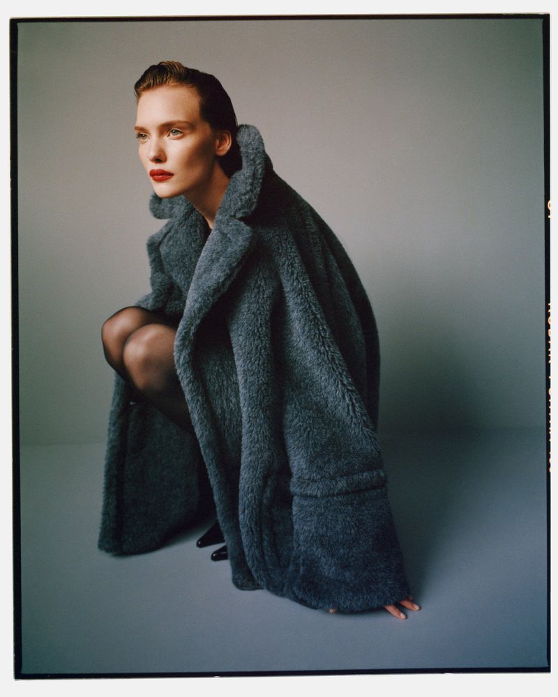 A woman with fair skin and dark hair wears a gray coat and sits in a squatting position on a gray background, creating a fashion editorial shot.