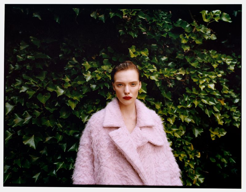 A fashionable woman with dark red lipstick poses outdoors, wearing a textured pink coat against a background of dark green ivy leaves.