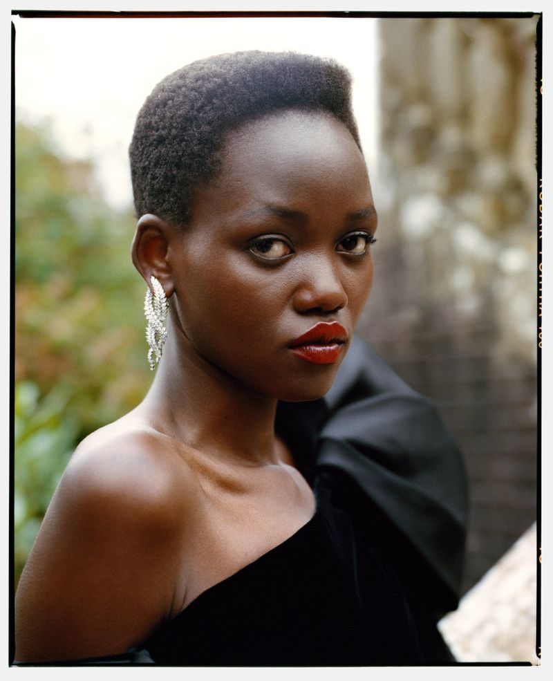 Close-up of elegant African-American woman in off-shoulder black velvet dress and crystal earrings against a muted outdoor background.