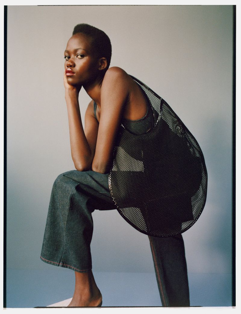 A dark-skinned woman poses indoors wearing a black mesh top and denim jeans, creating a fashion-forward, portrait-style studio shot.