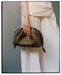A model in a draped off-white outfit holds an olive green leather drawstring clutch bag against a neutral studio backdrop.