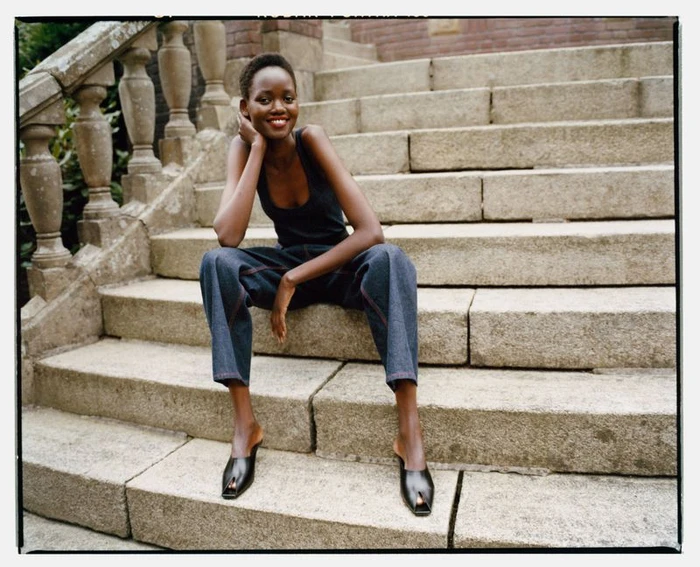 Model Caren Jepkemei smiling on outdoor stone steps in a black sleeveless top, wide-leg denim jeans, and black open-toe mules.