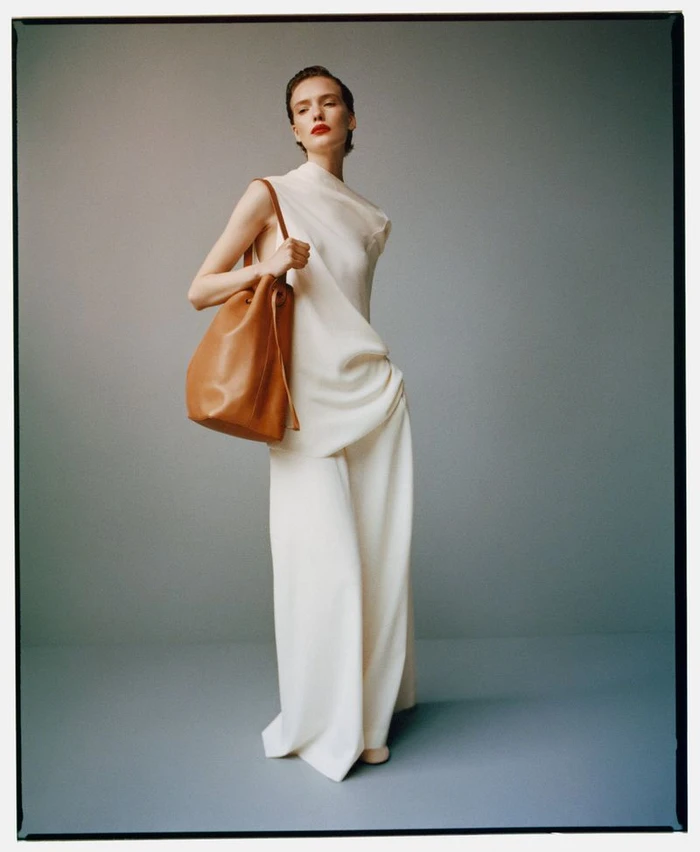Model Penelope Ternes in an off-white draped top and wide-leg trousers holding a brown leather shoulder bag against a grey backdrop.