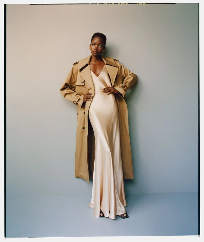 Model Caren Jepkemei in an open tan trench coat over a floor-length champagne satin slip dress against a pale grey seamless background.