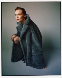 Model Penelope Ternes crouching in an oversized slate grey shearling coat and black tights against a grey studio backdrop.