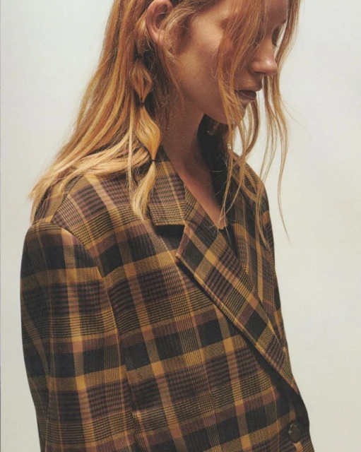 A woman with blonde hair in pigtails is wearing a brown plaid blazer against a white background, shot in a medium studio shot.