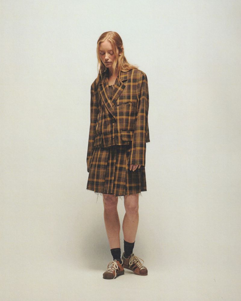 Young adult woman poses in a plaid blazer and pleated mini skirt, complemented by striped sneakers, against a neutral backdrop in a fashion editorial.