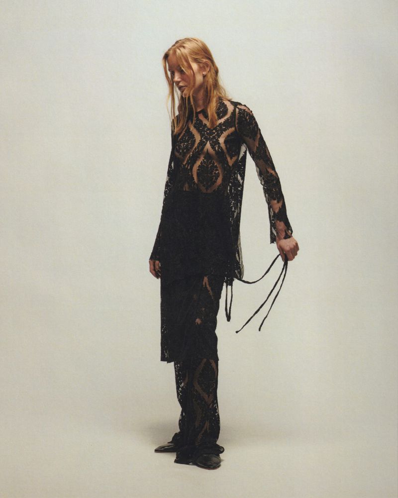 A slender woman with long blonde hair poses indoors against a pale backdrop, wearing a flowing black lace top and trousers.