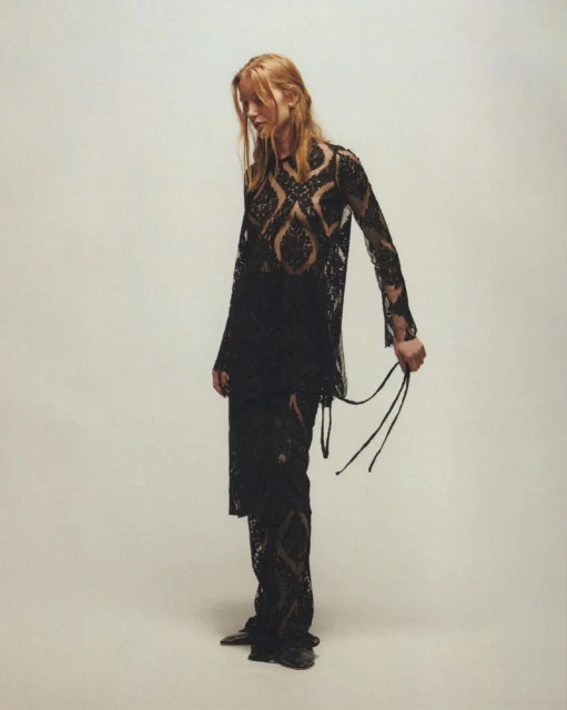 A slender woman with long blonde hair poses indoors against a pale backdrop, wearing a flowing black lace top and trousers.