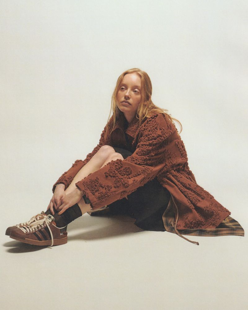A red-haired woman sits cross-legged on a cream background, wearing a brown coat and brown Adidas sneakers, with zebra stripes.