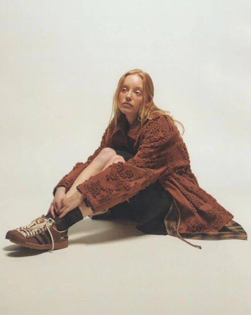 A red-haired woman sits cross-legged on a cream background, wearing a brown coat and brown Adidas sneakers, with zebra stripes.
