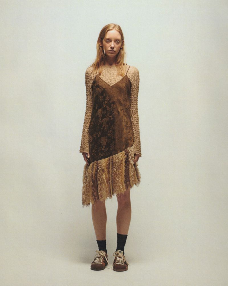 Jeske van der Pal wearing a layered brown fringe slip dress with knit sleeves and sneakers against a plain grey studio backdrop.