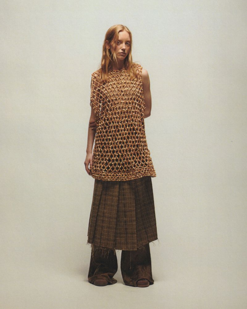 A single young adult female model stands indoors wearing a brown knit top and a long brown plaid skirt, against an off-white backdrop.