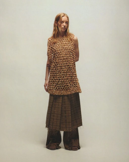 A single young adult female model stands indoors wearing a brown knit top and a long brown plaid skirt, against an off-white backdrop.