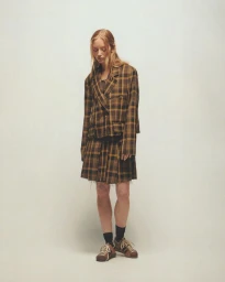 Model standing on a plain light background wearing a plaid blazer and matching pleated skirt with sneakers