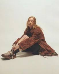 Model wearing a textured brown coat and brown sneakers sitting against a plain white background