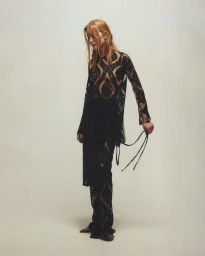 Model Jeske van der Pal wearing a sheer patterned black tunic top and matching trousers against a pale studio seamless backdrop.