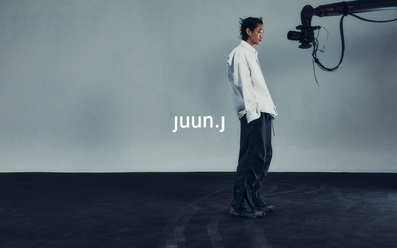 Fashion shot of a slender adult male in a white shirt and dark pants, standing in a studio near film equipment. 