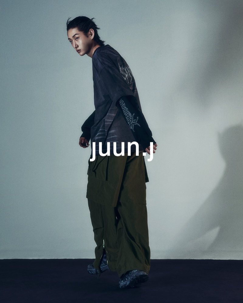 A young Asian man poses indoors wearing a black t-shirt, olive cargo pants, and sneakers, showcasing a casual yet stylish fashion ensemble.