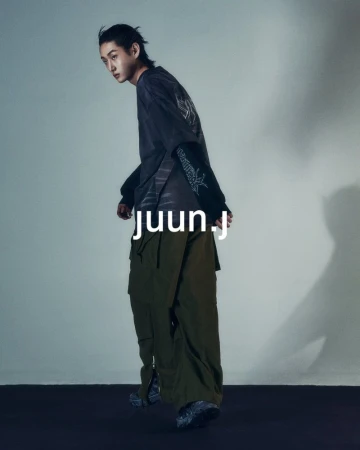 A young Asian man poses indoors wearing a black t-shirt, olive cargo pants, and sneakers, showcasing a casual yet stylish fashion ensemble.