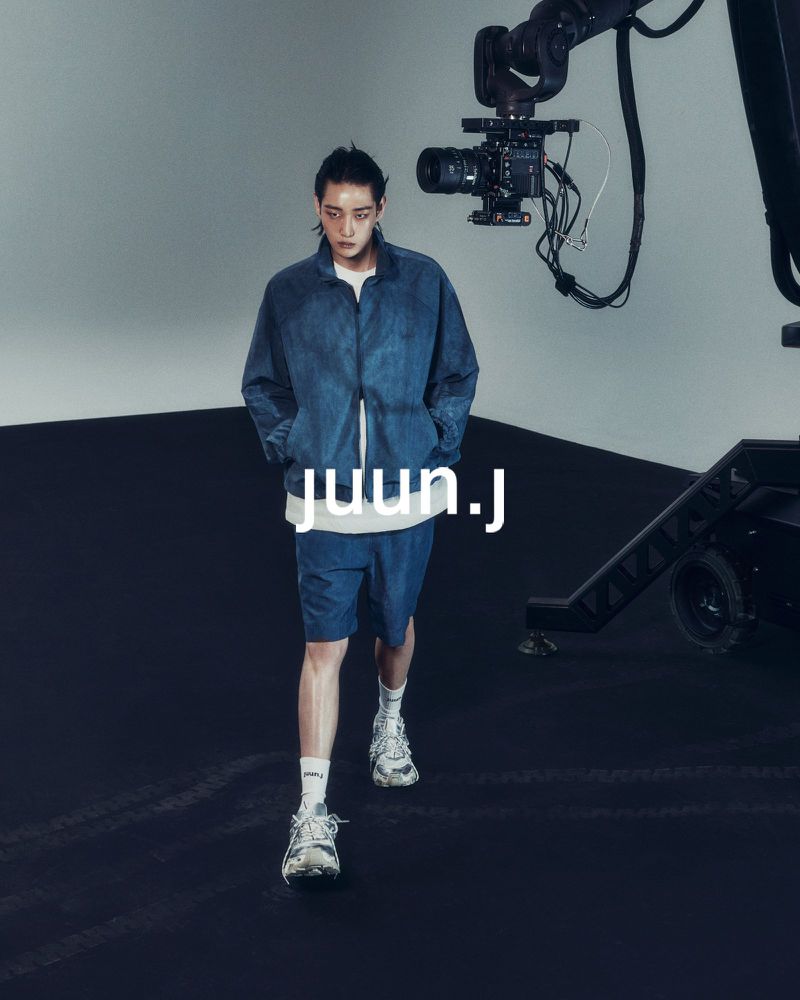 Male fashion model in blue denim jacket and shorts walks toward the camera in a studio setting with robotic camera arm in the background.