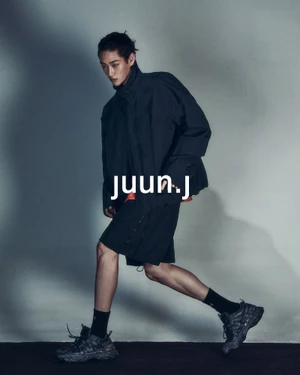 Model in dark jacket and shorts walking against a grey studio seamless backdrop with brand logo