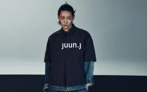 Model Hamin Yu wears a layered Juun.J black short-sleeve shirt over denim sleeves against a cool grey studio backdrop.