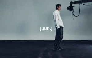 Model Hamin Yu standing in profile wearing an oversized white shirt and wide trousers next to a studio camera rig with the Juun.J logo.