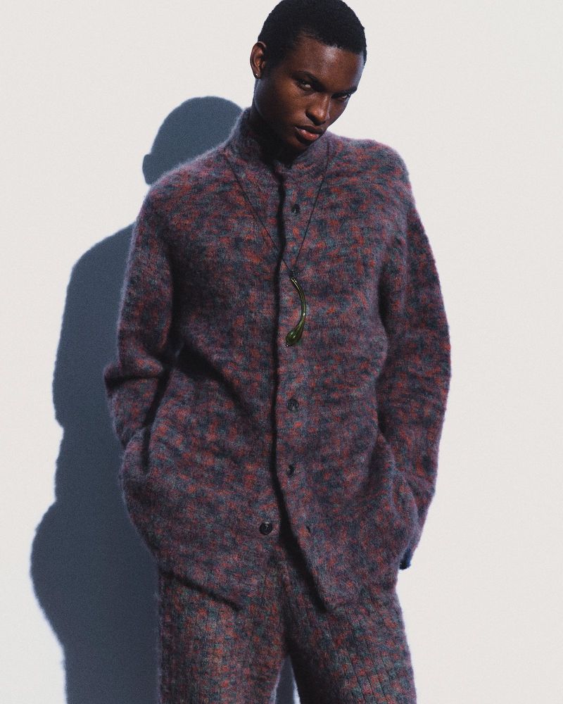 An androgynous person wearing a textured purple and red sweater and matching pants poses in front of a white wall with their shadow.