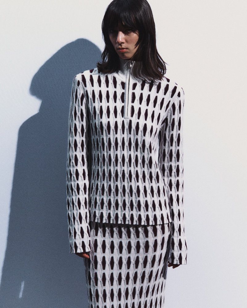 Model wearing a black and white geometric knit zip-up sweater and matching skirt against a white studio backdrop with a cast shadow.