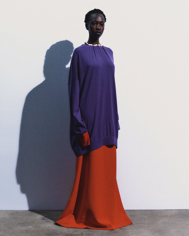 A tall, dark-skinned fashion model stands in a photo studio wearing a purple top and a red skirt against a white backdrop and shadow.