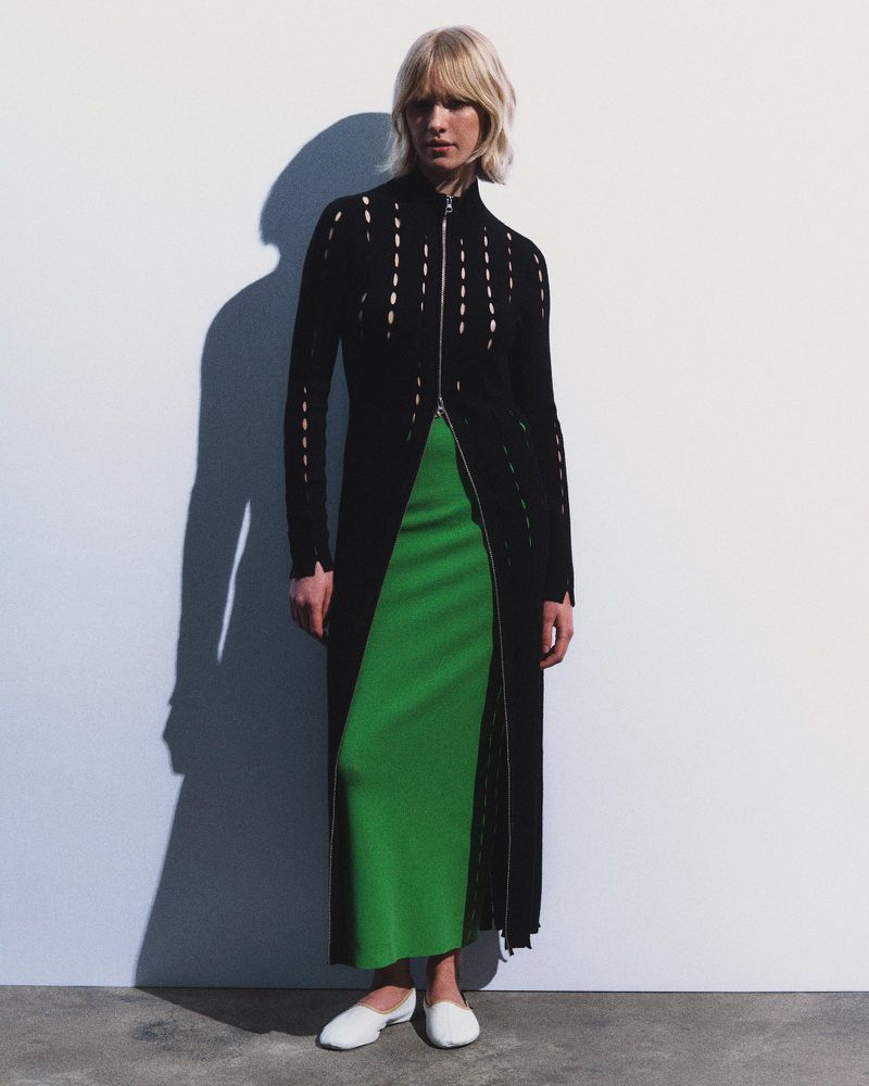 Fashionable woman with blond hair poses in a black coat over a green skirt, standing in a photo studio with a white backdrop.