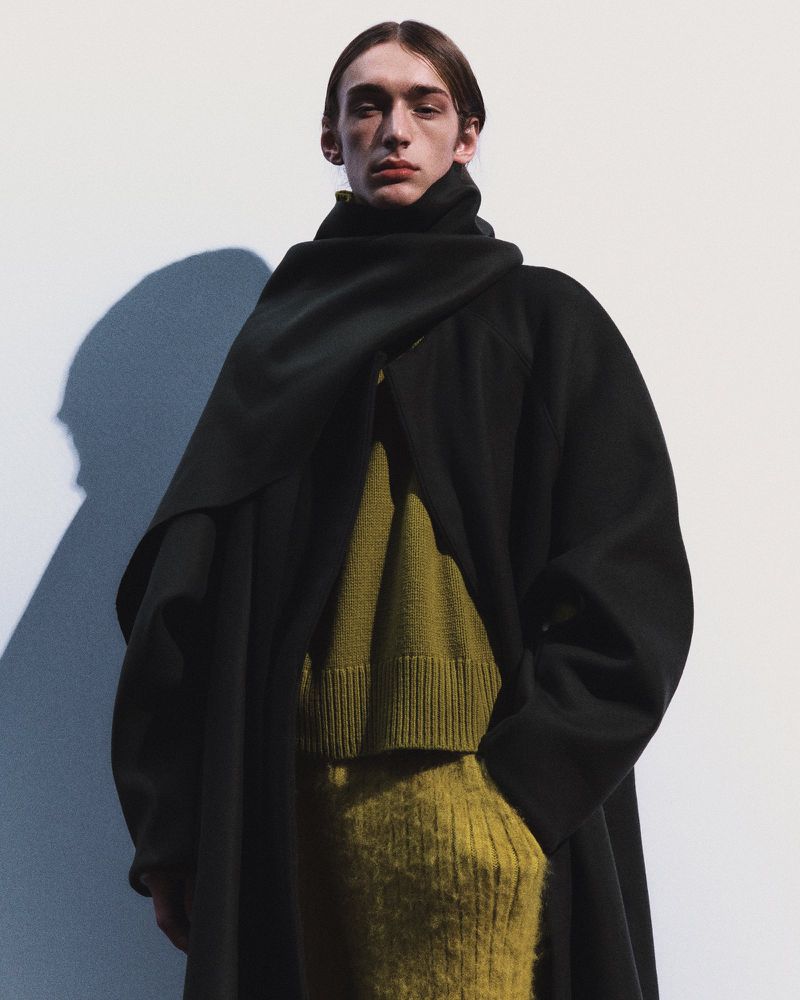 Model Julien Saunier wearing an oversized black coat with draped scarf collar over an olive green knit sweater against a white backdrop.