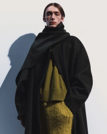 Model Julien Saunier wearing an oversized black coat with draped scarf collar over an olive green knit sweater against a white backdrop.